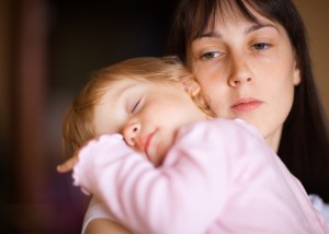 A sad-looking young mother holding a sleeping toddler against her shoulder